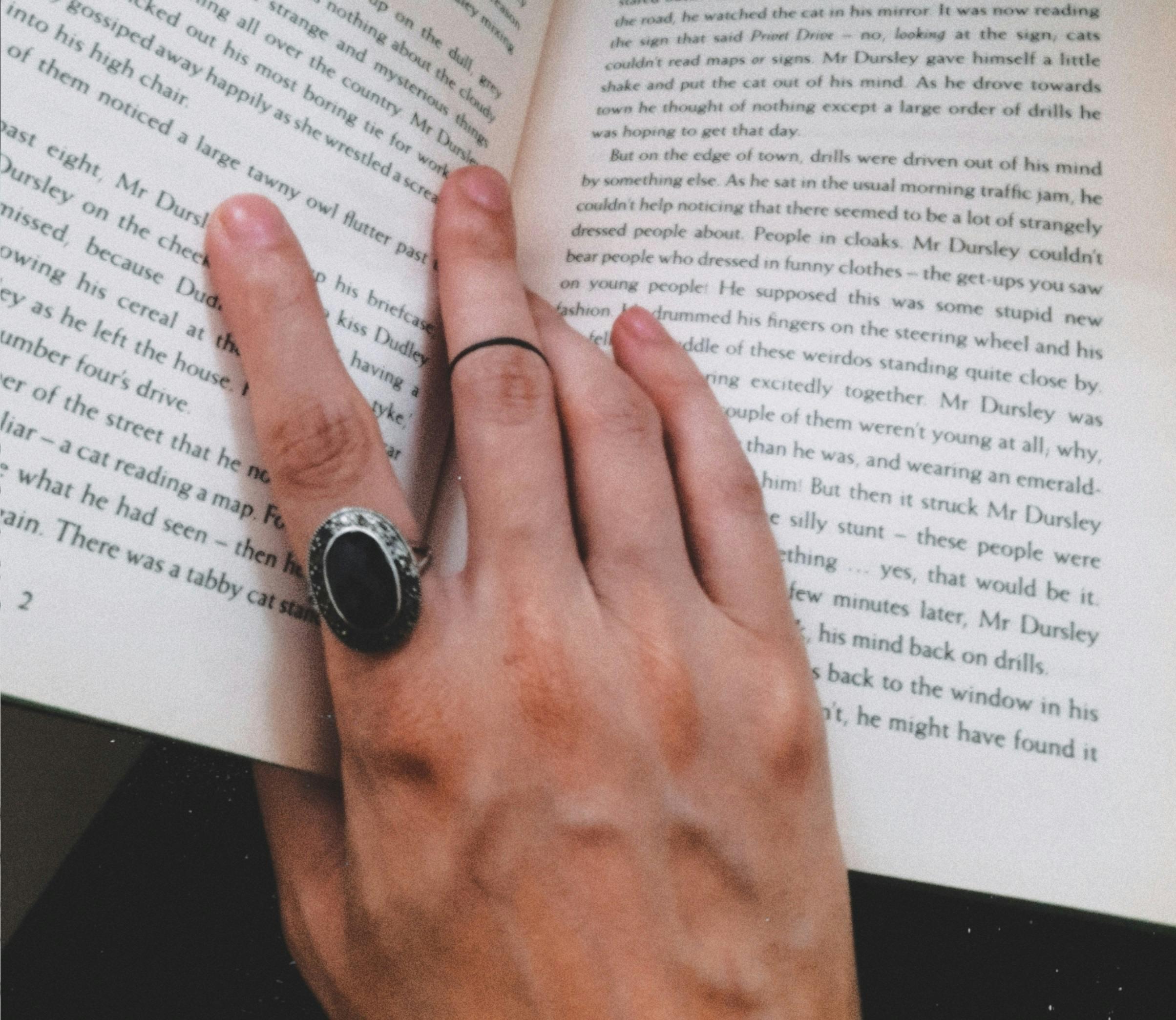 Na foto há uma mão esticada sobre um livro aberto, a mão é branca e usa um anel preto no indicador.